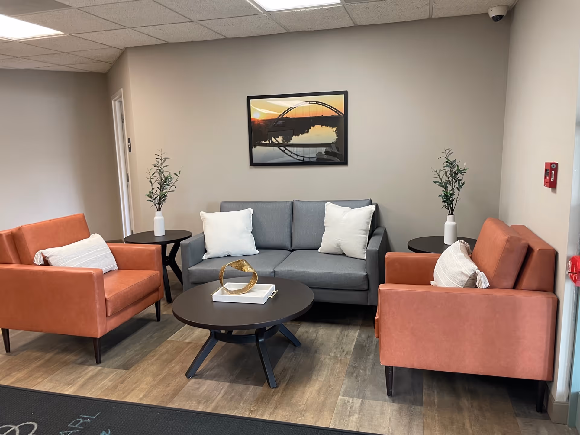 A small lobby seating area with a gray sofa, two orange armchairs, round black tables, decorative pillows and wall art.