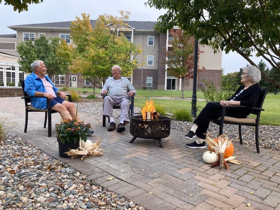 Three elderly people sitting outdoors on chairs around a fire pit on a paved patio. There are decorative pumpkins and plants near the chairs. In the background, there is a multi-story senior living apartment building with trees and grass.