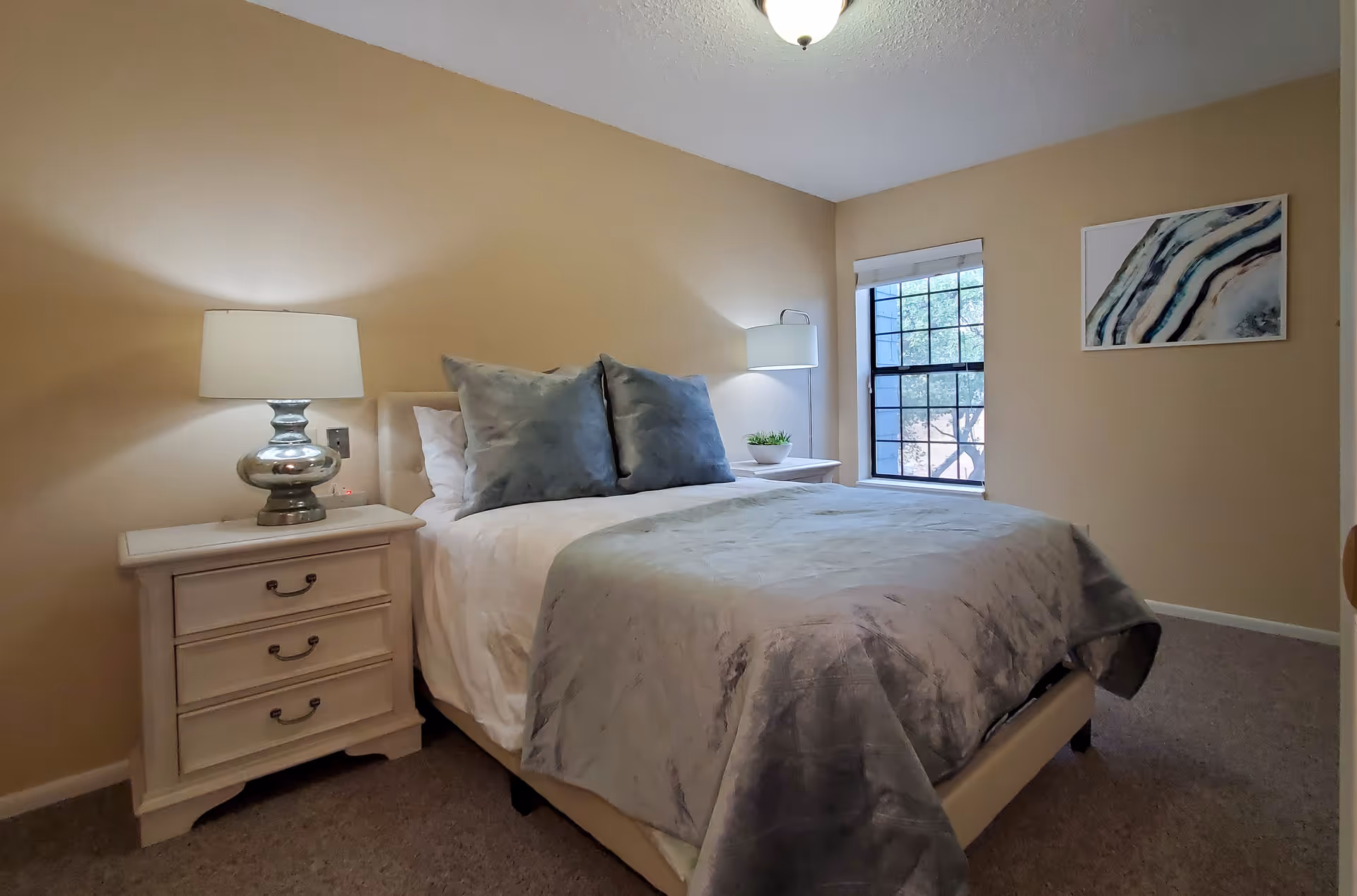 A cozy bedroom with beige walls featuring a bed with white sheets, a gray blanket, and two large gray pillows. There are two white nightstands on either side of the bed, each with a lamp. A window with a white blind is on the right wall, and a framed abstract artwork hangs next to it.