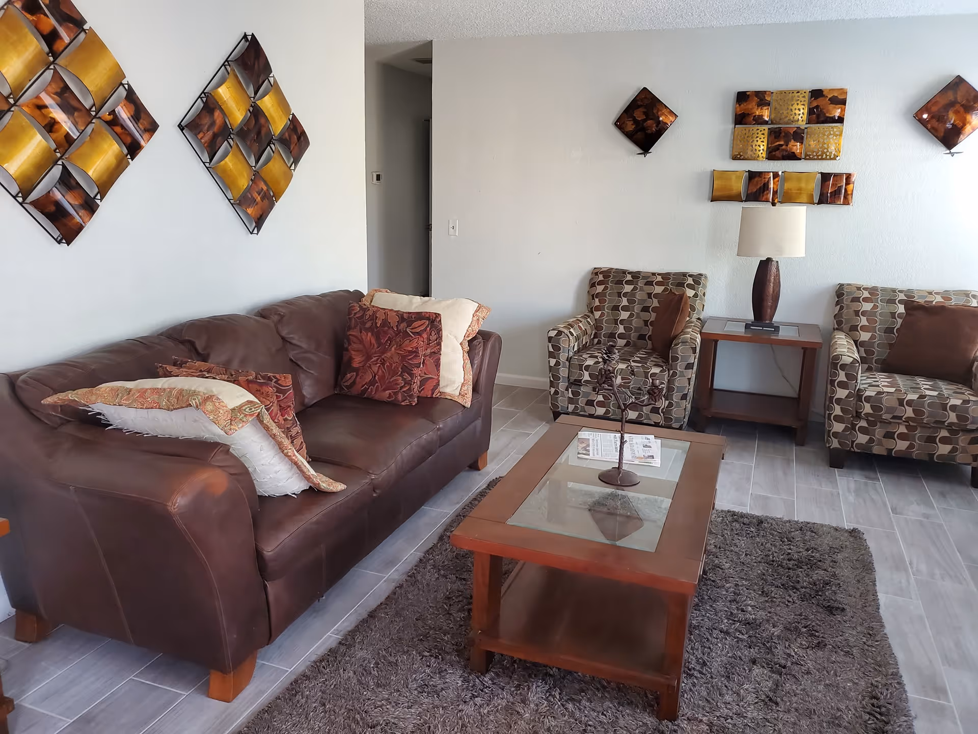 A cozy living room with a brown leather sofa adorned with multiple patterned cushions, two patterned armchairs with brown pillows, a wooden coffee table with a glass top and decorative centerpiece, a side table with a lamp, and abstract wall art in shades of brown and gold on a white wall. The floor is tiled with a gray rug in front of the sofa.