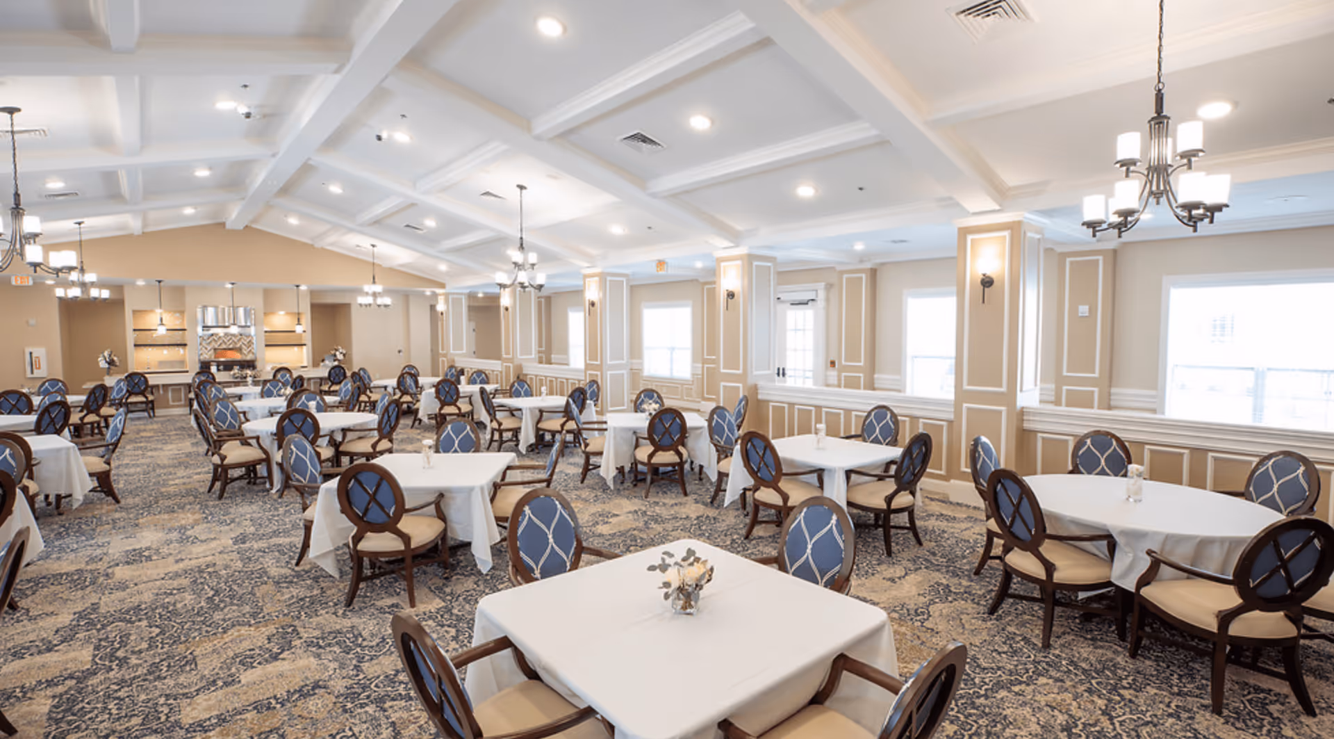 A spacious, well-lit dining room with multiple round and square tables covered with white tablecloths. Each table is surrounded by elegant wooden chairs with blue cushioned seats and backs featuring a diamond pattern. The room has a high coffered ceiling with recessed lighting and several chandeliers. The walls are beige with white trim, and large windows allow natural light to fill the space. The carpet has a blue and beige intricate pattern.