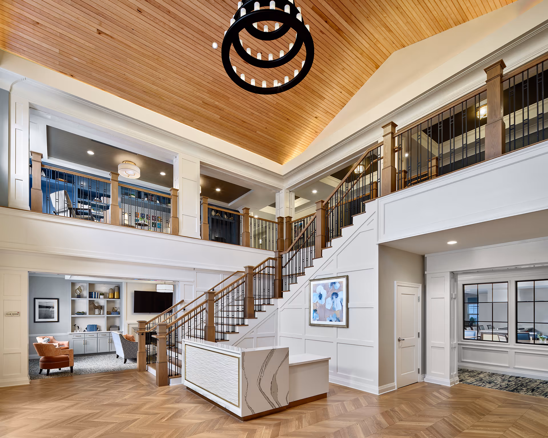 Spacious and elegant interior of a senior living facility featuring a large wooden staircase with black metal railings, a high wooden ceiling with a modern circular chandelier, and a reception desk with a marble-like finish. The area includes a seating space with chairs and a TV, and a hallway leading to other rooms.