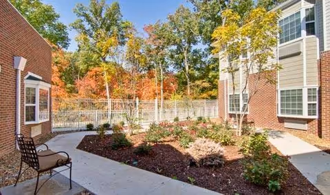 Outdoor courtyard area between two brick buildings with a concrete pathway, garden beds with various plants and shrubs, and a metal chair with a cushion. Trees with autumn-colored leaves are visible in the background.