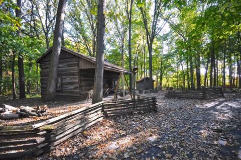 A wooded outdoor area with tall trees and sunlight filtering through the leaves. There are rustic wooden cabins and split-rail fences surrounded by fallen leaves on the ground.