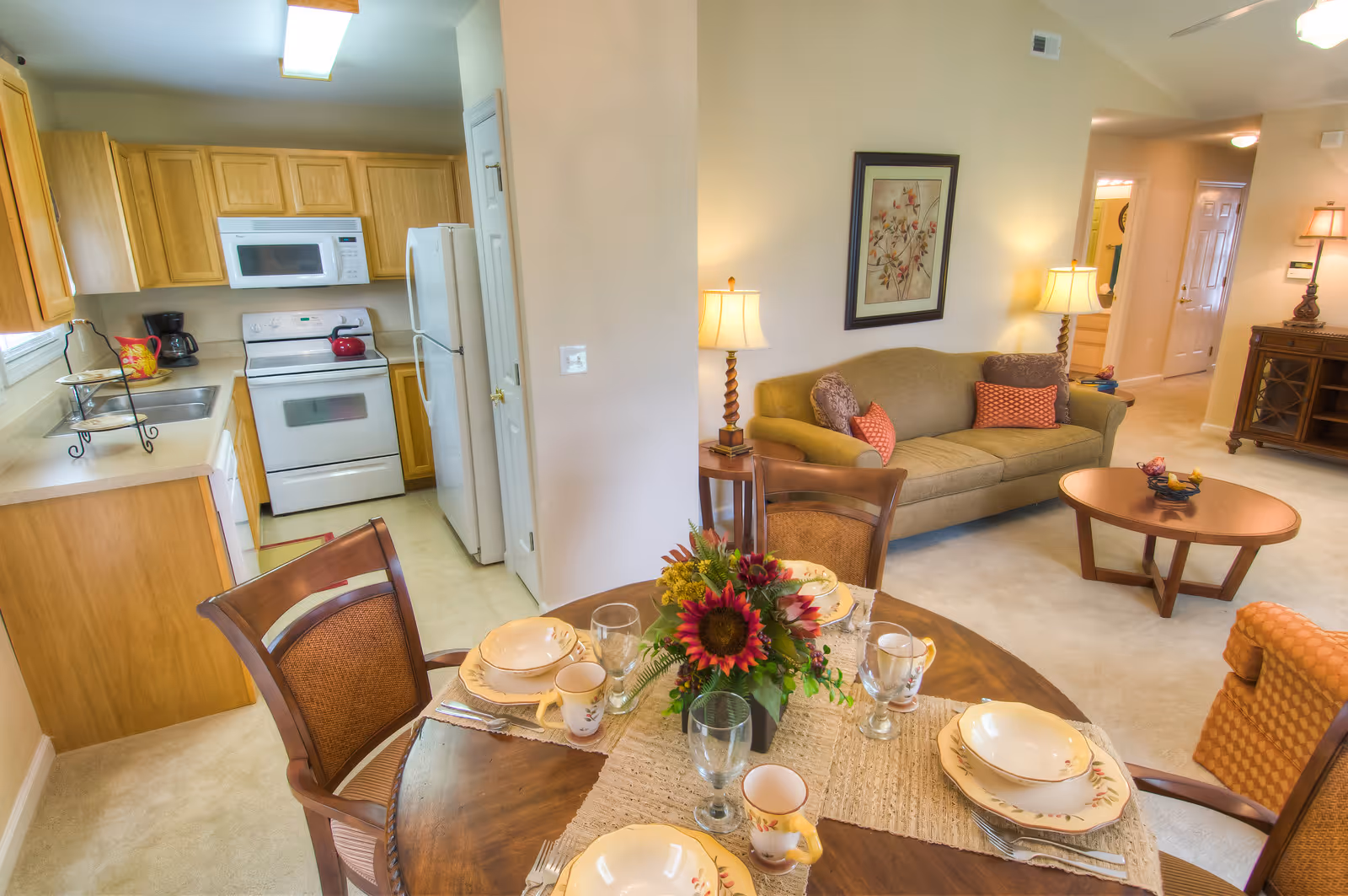 View of a cozy senior living apartment featuring a small kitchen with wooden cabinets, white appliances including a stove, microwave, and refrigerator. Adjacent to the kitchen is a dining area with a round wooden table set with plates, bowls, mugs, glasses, and a floral centerpiece. Beyond the dining area is a living room with a beige sofa adorned with decorative pillows, a wooden coffee table, side tables with lamps, and framed artwork on the wall. The space is warmly lit and carpeted.