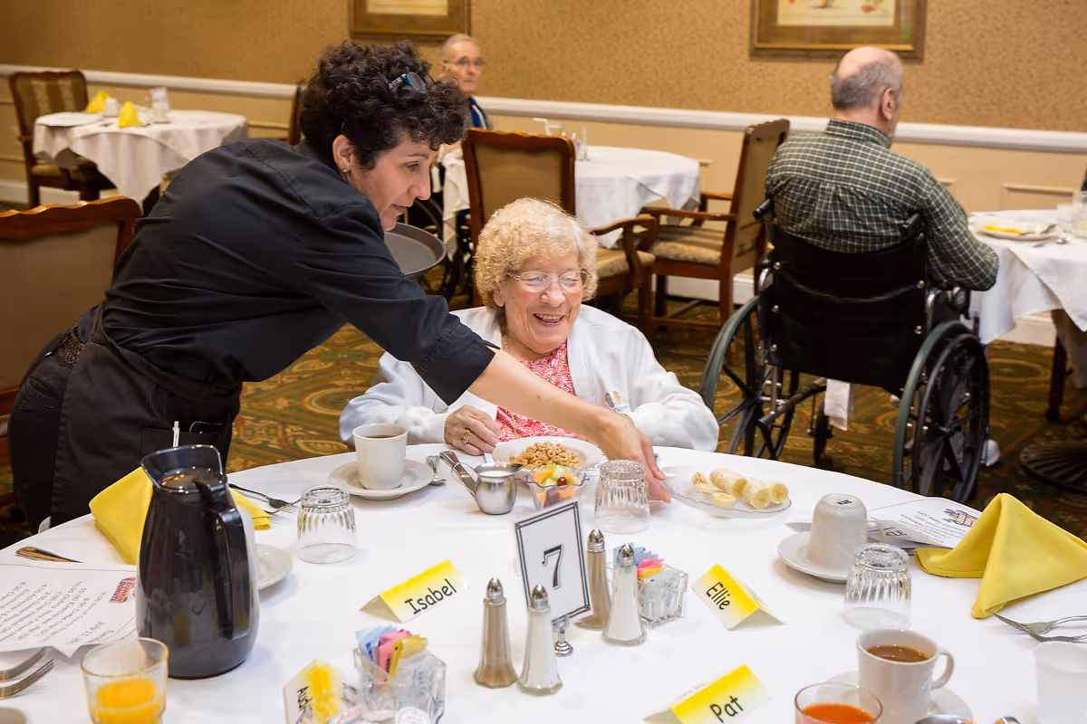 A dining room scene at Independence Village of Aurora where a caregiver is serving a plate of sliced bananas to a smiling elderly woman seated at a table. The table is set with breakfast items including cereal, coffee, juice, and place cards with names. Other elderly residents are visible in the background, some seated in wheelchairs.