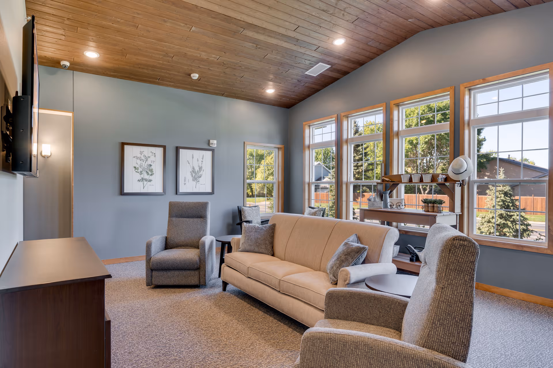 Bright lounge with a beige sofa and upholstered armchairs facing a wall-mounted TV, large windows, and a wood-paneled ceiling.