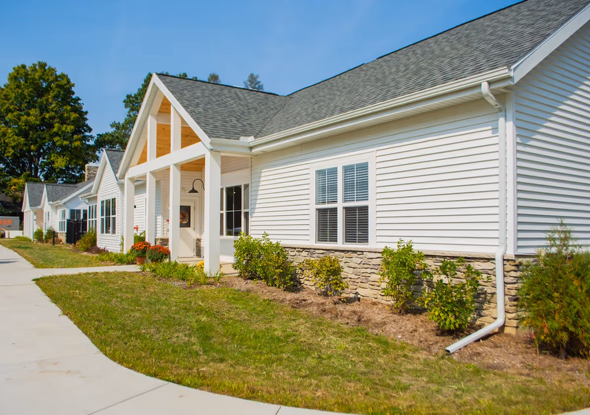 Exterior view of a single-story building with white siding and stone accents, featuring a covered entrance with white pillars and a shingled roof. There are bushes and plants along the building and a concrete walkway in front under a clear blue sky.