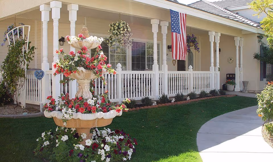 Front porch of a senior care facility with white railing and columns, an American flag hanging, and a tiered flower fountain with red, white, and purple flowers in the foreground. The area is landscaped with green grass and bushes.