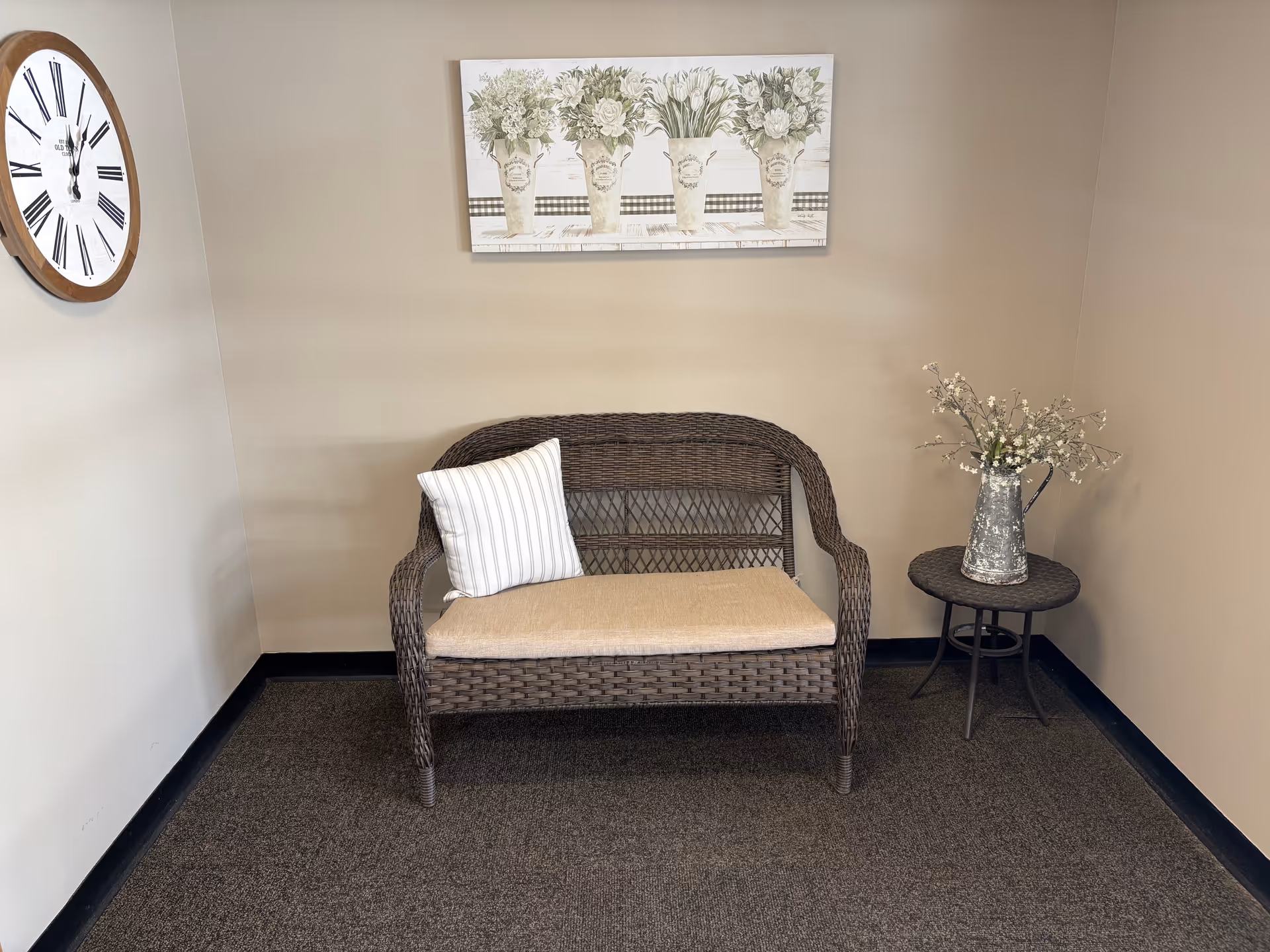 A small seating area with a wicker loveseat featuring a beige cushion and a white striped pillow. To the right of the loveseat is a round wicker side table with a metal pitcher holding white flowers. On the wall above the loveseat is a painting of four vases filled with white flowers. A large round wall clock with Roman numerals is mounted on the left wall. The walls are painted beige and the floor is carpeted in a dark color.