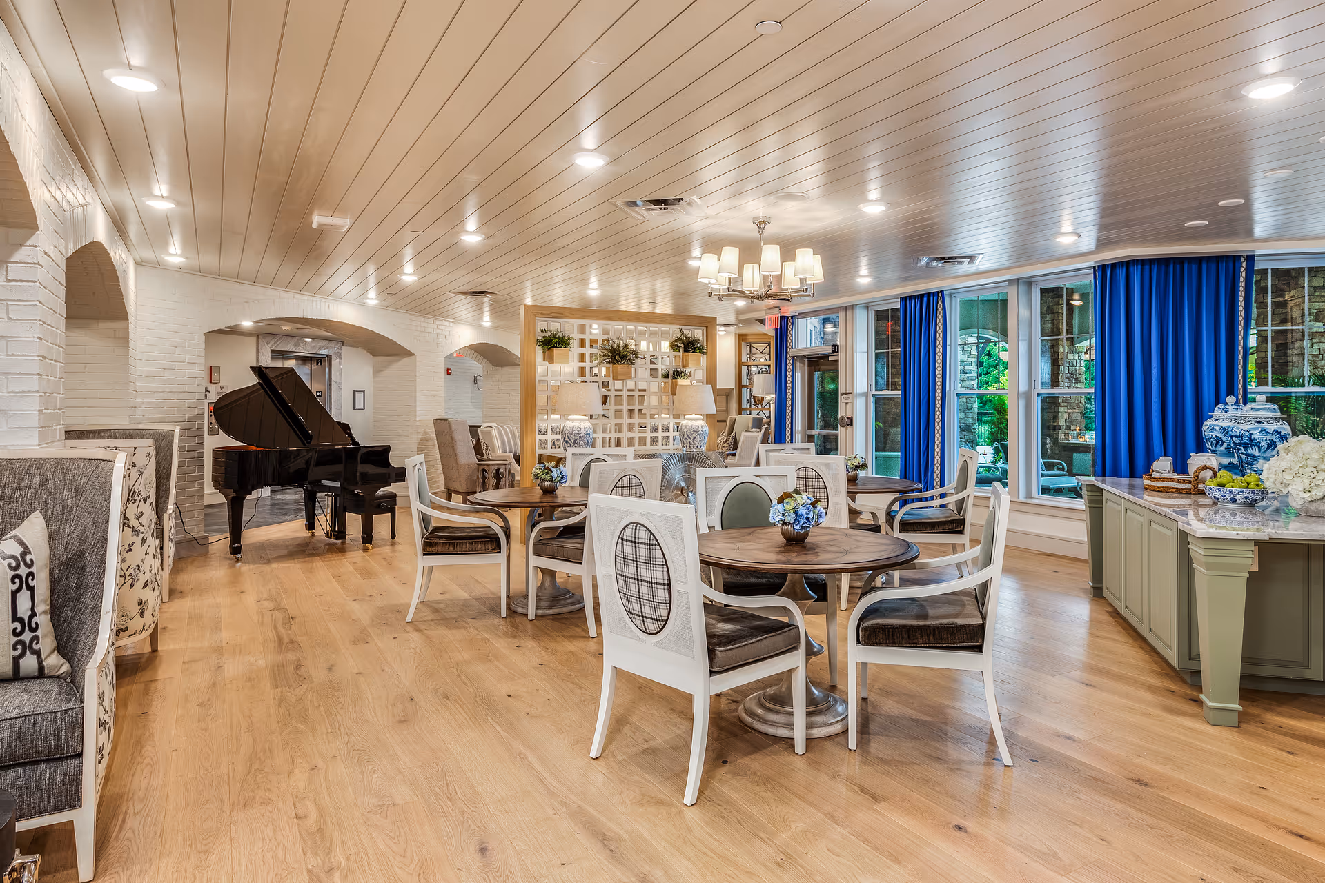 A spacious and well-lit common area with wooden floors and a white paneled ceiling. The room features several round wooden tables with white chairs, a black grand piano in the corner, blue curtains on large windows, and decorative plants and vases. There is a chandelier hanging from the ceiling and a green cabinet with decorative items on the right side.