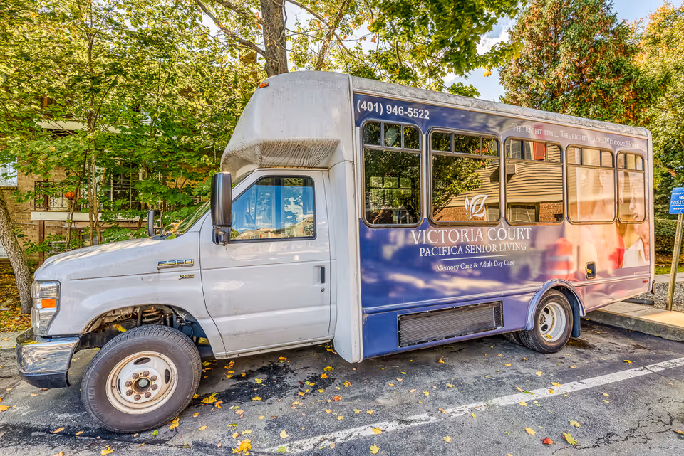 A white and blue shuttle bus parked in a lot surrounded by trees and a building. The bus has signage for Victoria Court Pacifica Senior Living, offering memory care and adult day care services, with a phone number (401) 946-5522 displayed on the side.