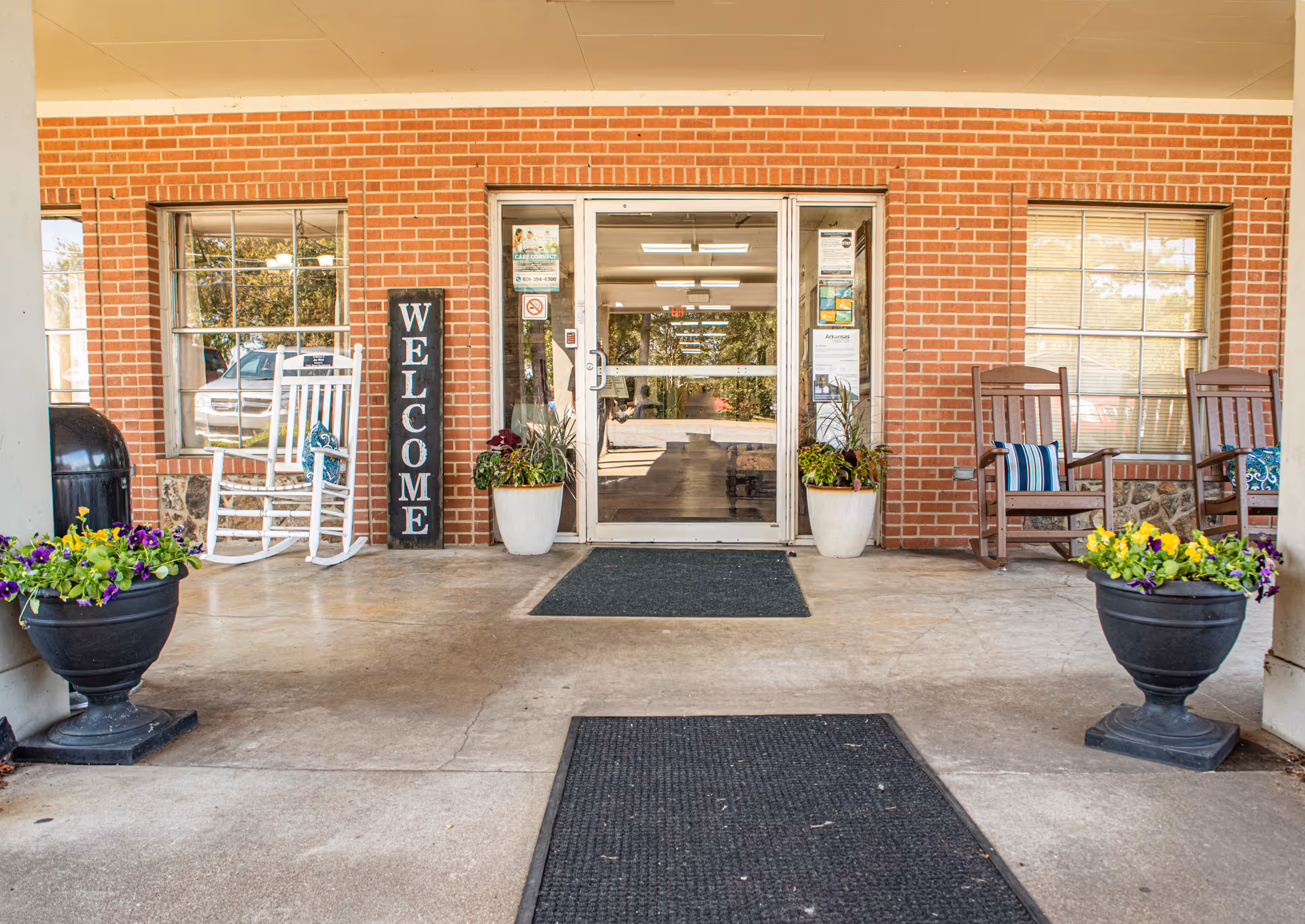 Covered brick entrance with glass double doors, a 'WELCOME' sign, potted flowers and rocking chairs.