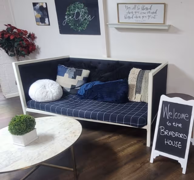 A cozy seating area with a modern cushioned bench adorned with various decorative pillows. A small round marble table with a potted plant sits in front of the bench. On the wall behind, there are framed decorative signs, one with the word 'gather' and another with a quote. A small chalkboard sign on the floor reads 'Welcome to the Bradford House'.