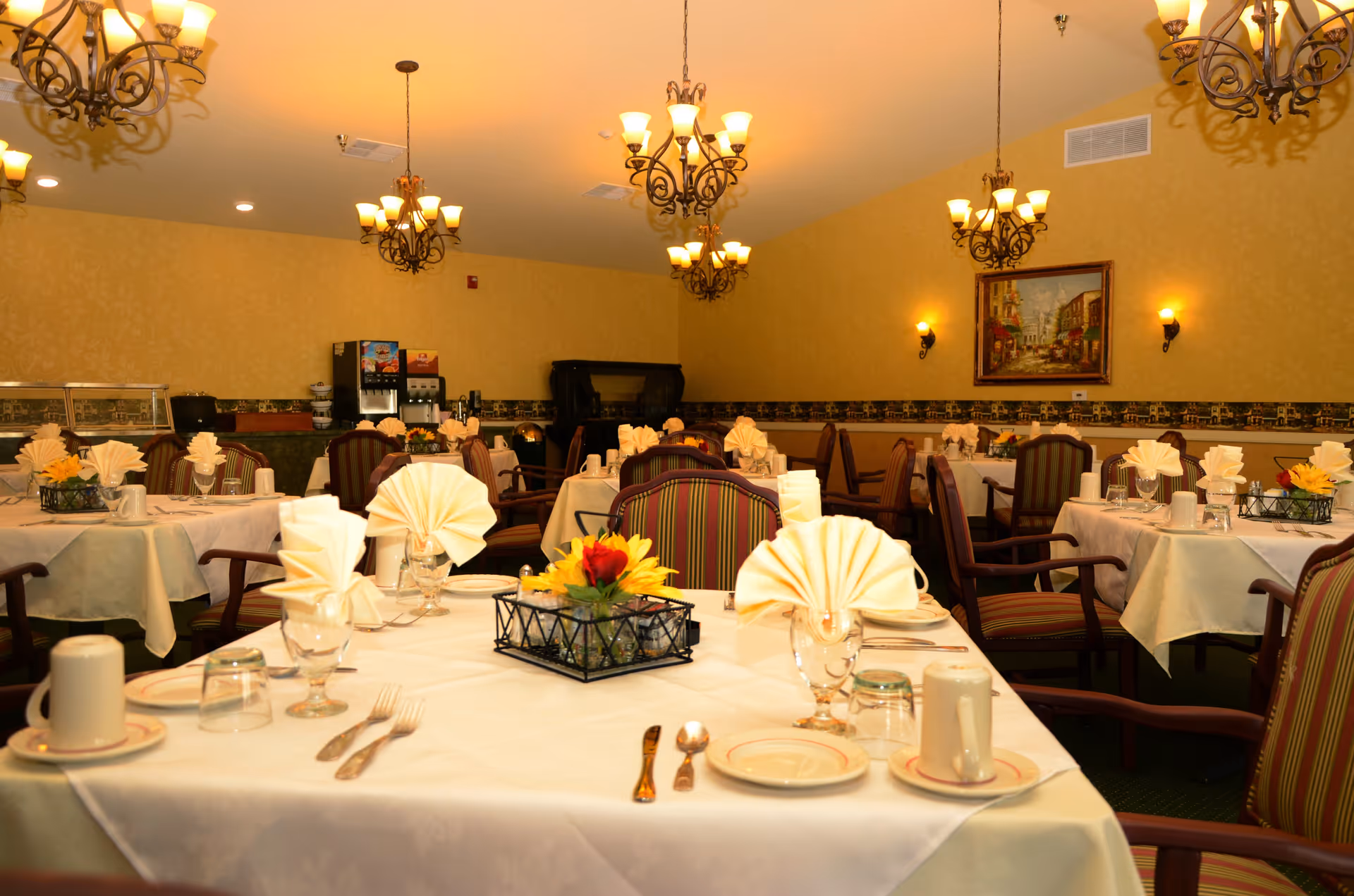 A warmly lit dining room with multiple tables covered in white tablecloths, each set with folded napkins, plates, glasses, and silverware. The chairs have striped upholstery. There are several chandeliers hanging from the ceiling, wall sconces providing additional light, and a framed painting on the wall. In the background, there is a beverage dispenser and a buffet area.