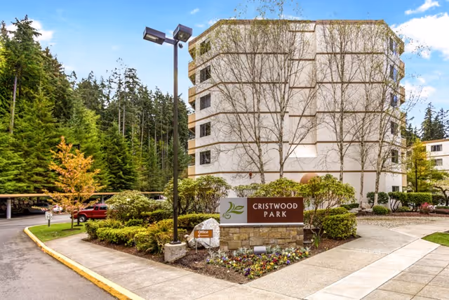 Exterior view of a multi-story residential building surrounded by trees and landscaped greenery. A sign in front reads 'Cristwood Park' with a small 'Office' sign nearby. There is a driveway and parking area with a red vehicle visible, and the sky is partly cloudy.