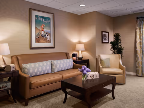 Cozy living room with brown leather sofa, armchair, coffee table, lamps, artwork, and plants.
