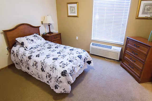 A bedroom with a single bed covered in a white and black floral patterned comforter. There is a wooden headboard, a wooden nightstand with a lamp, a wooden dresser, two framed pictures on the walls, a window with closed blinds, and a wall-mounted air conditioning unit below the window.