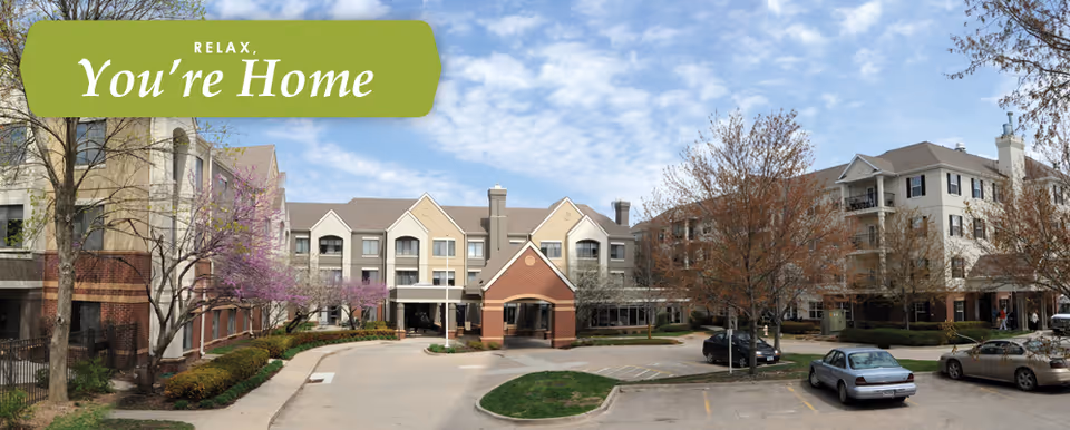 Front exterior of a multi-story senior living campus with a covered entrance, parking lot, and landscaped trees under a blue sky.
