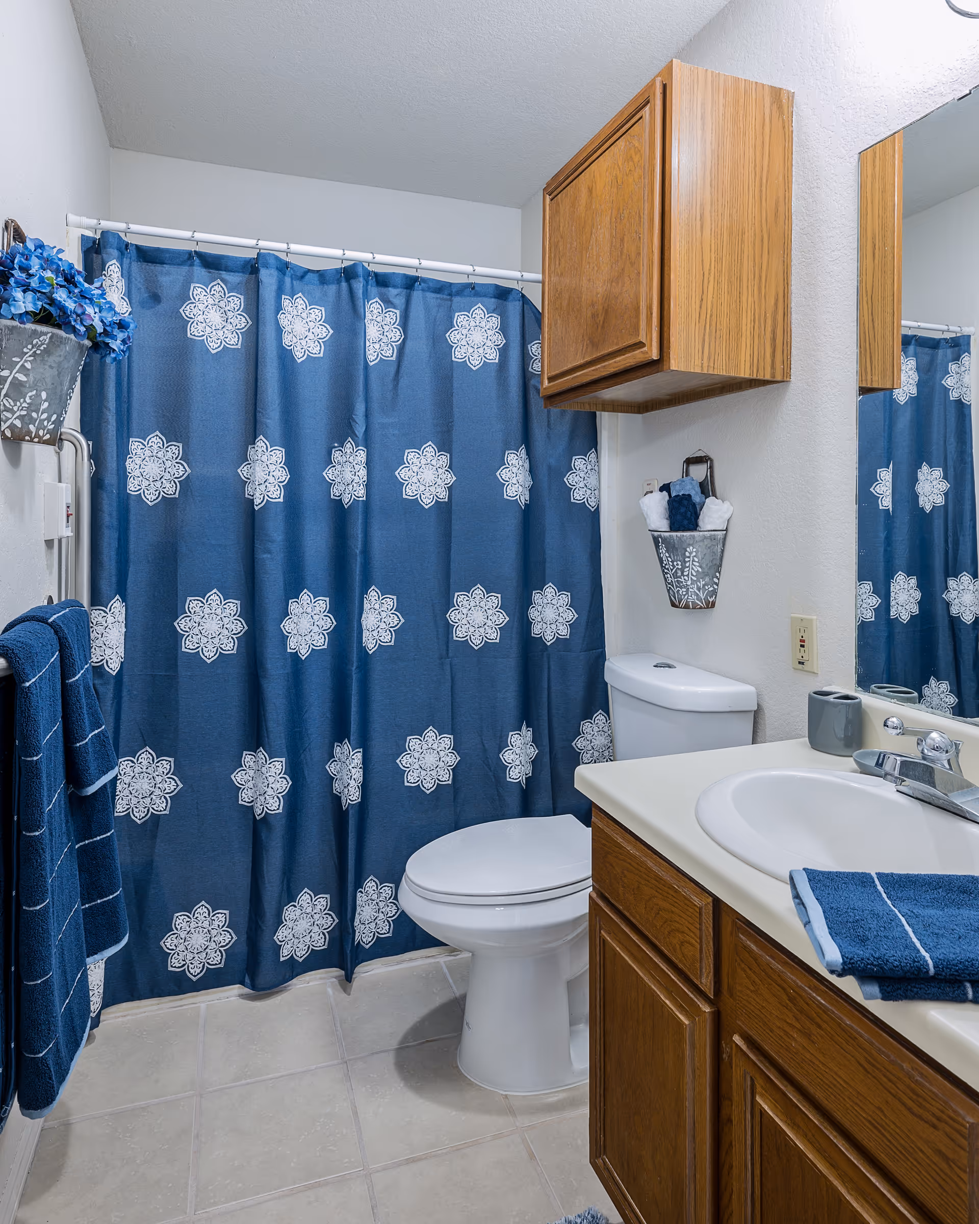 A bathroom with a white toilet, a wooden cabinet above it, and a white sink with a wooden vanity. There is a blue shower curtain with white floral patterns, blue towels hanging on a rack, and a small metal container holding rolled towels on the wall. The floor is tiled in light beige.