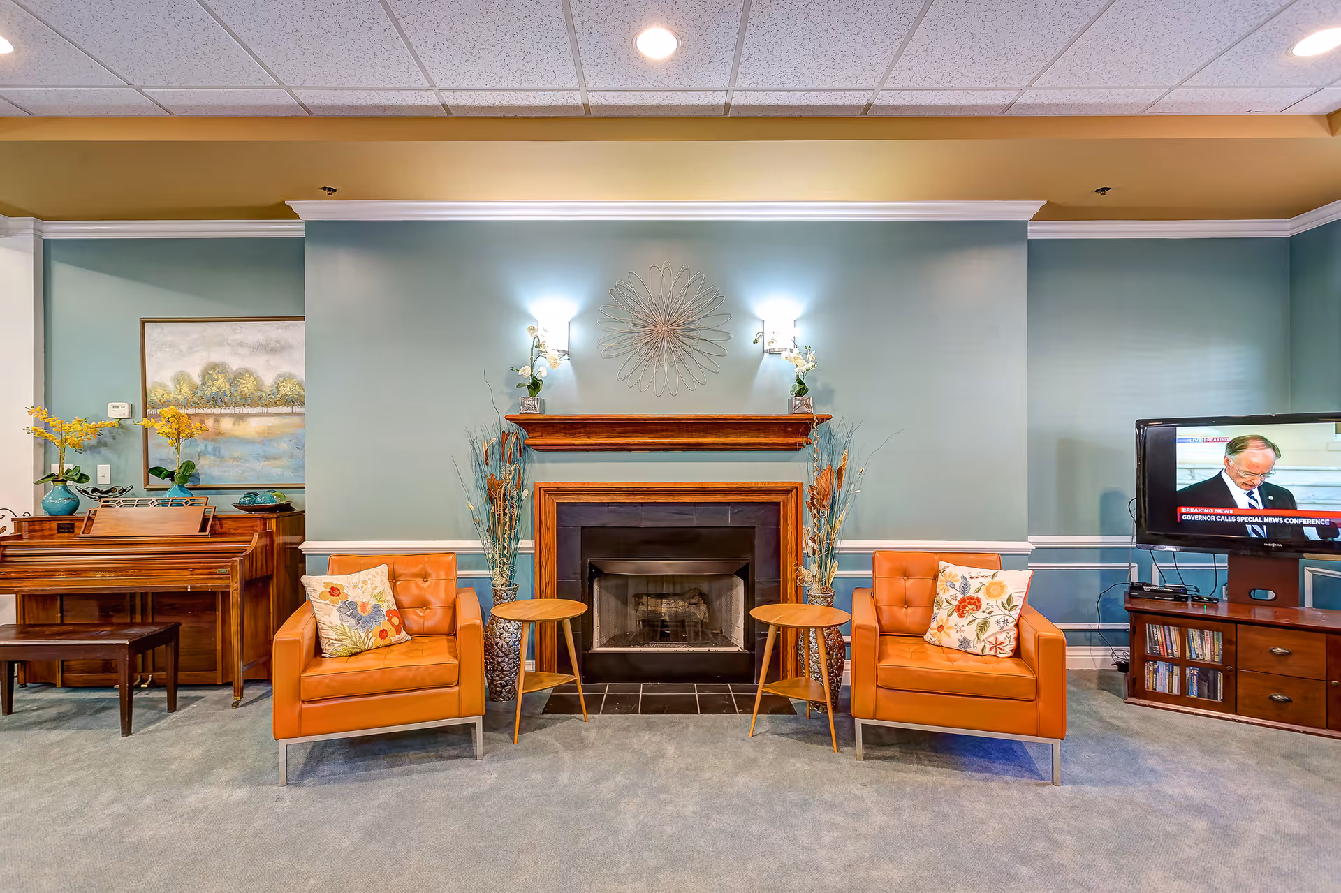 Comfortable lounge area with a central fireplace flanked by two orange armchairs, side tables, a piano to the left and a TV to the right.