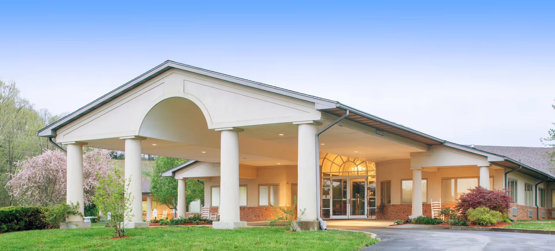 Front entrance of a single-story senior living facility with a covered porte-cochere, columns, glass doors, and landscaped lawn.