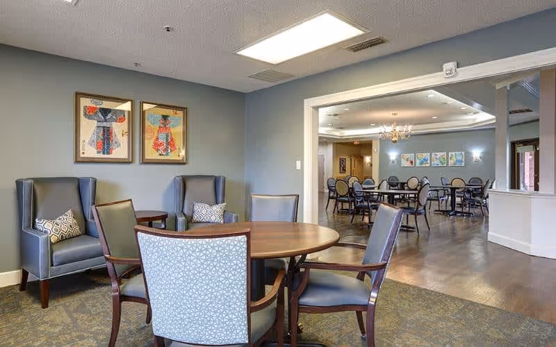 A seating area in a senior living facility with a round wooden table surrounded by four chairs with patterned upholstery. Two gray armchairs with decorative pillows are positioned against a blue-gray wall with two framed artworks. In the background, there is a larger dining area with multiple tables and chairs, wall art, and a chandelier.