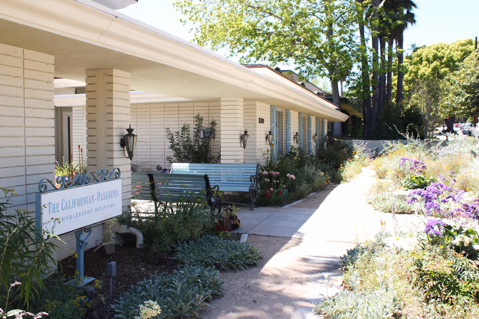Front entrance of The Californian - Pasadena convalescent hospital with benches and landscaped flower beds along a sunny walkway.