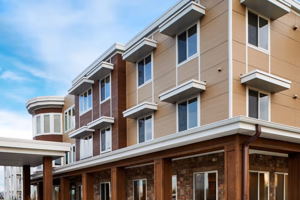 Exterior view of a modern three-story senior living facility building with beige and brown paneling, multiple windows, and a covered entrance under a partly cloudy sky.