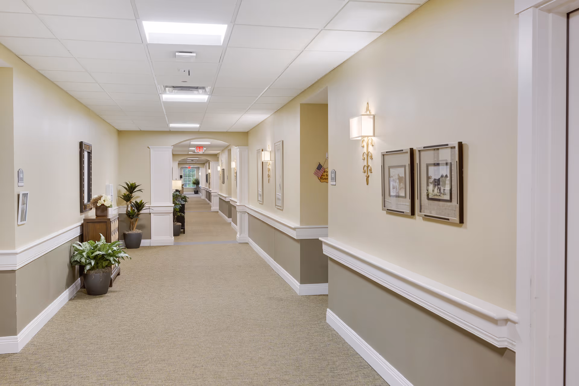 A long, well-lit hallway in a senior living facility with beige walls and carpeted floor. The hallway is decorated with framed pictures, wall sconces, and several potted plants. There is an exit sign visible at the far end of the corridor.