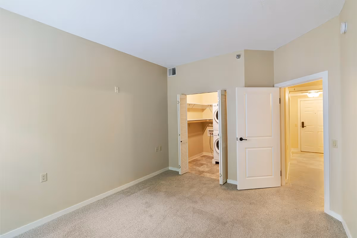 Empty room with beige walls and carpeted floor, featuring an open closet with a stacked washer and dryer inside, and an open door leading to a hallway with a white entrance door visible.