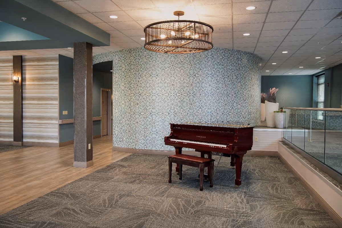 Interior view of a senior living facility featuring a polished wooden piano with a matching bench placed on a patterned carpet. The background includes a curved wall with intricate blue and white tile design, a modern circular ceiling light fixture, and a glass railing on the right side. The floor transitions from carpet to wood, and there are decorative plants in white pots near the window.