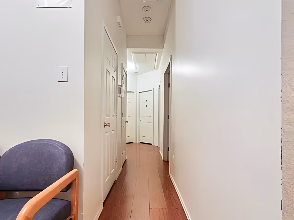 A narrow hallway with wooden flooring and white walls, featuring several closed white doors along the corridor. A blue cushioned chair with wooden armrests is partially visible on the left side.