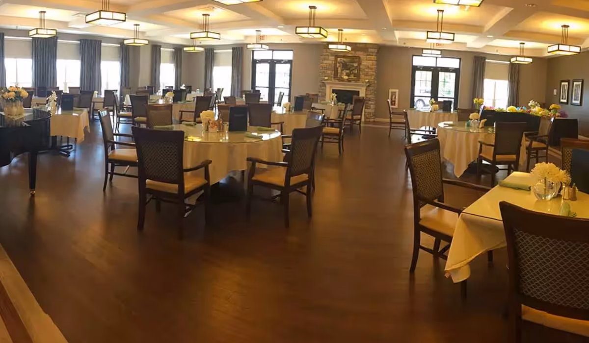 A spacious dining room with multiple round tables covered with beige tablecloths, each surrounded by wooden chairs with cushioned seats and backs. The room features large windows with curtains, a stone fireplace, and modern ceiling lights. There are floral centerpieces on the tables and a grand piano on the left side.
