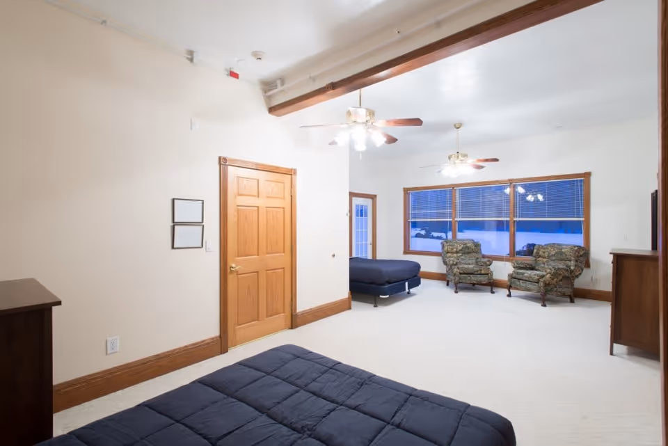 A spacious bedroom with two beds covered in dark bedding, two floral upholstered armchairs near large windows, wooden door and trim, ceiling fans with lights, and light-colored carpet.