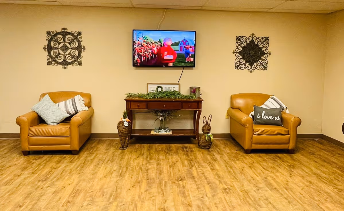 A cozy seating area with two tan leather armchairs, each adorned with decorative pillows. Between the chairs is a wooden console table with greenery and decorative items. Above the table, a flat-screen TV is mounted on the beige wall, displaying a colorful animated scene. Two ornate metal wall decorations flank the TV on either side. The floor is a light wood laminate.