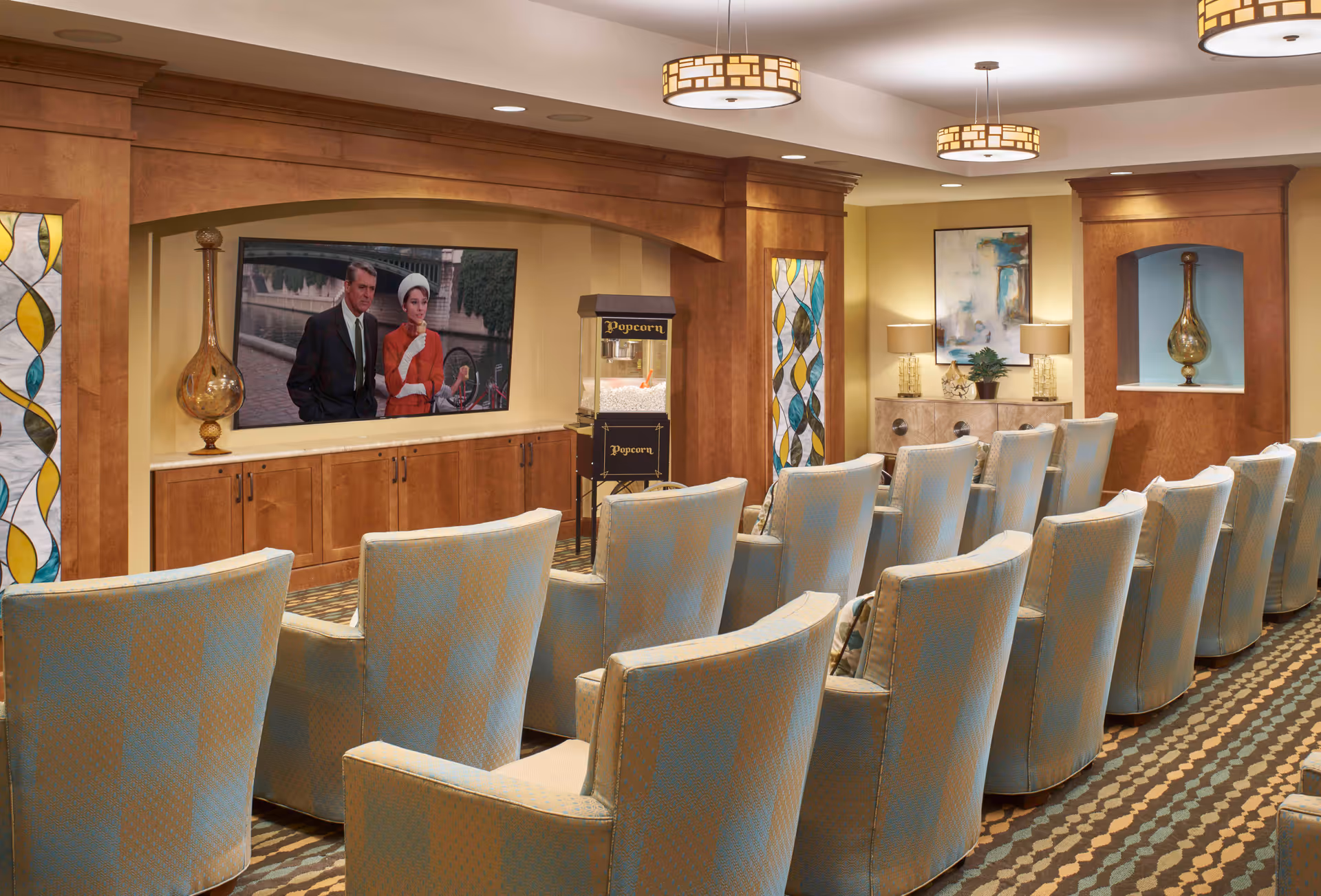 A cozy senior living facility theater room with two rows of comfortable upholstered chairs facing a large flat-screen TV mounted on a wooden wall. The TV is showing a classic movie scene with a man and woman. There is a popcorn machine next to the TV, decorative stained glass panels on the walls, and warm lighting from ceiling fixtures and table lamps on a sideboard with artwork and plants.