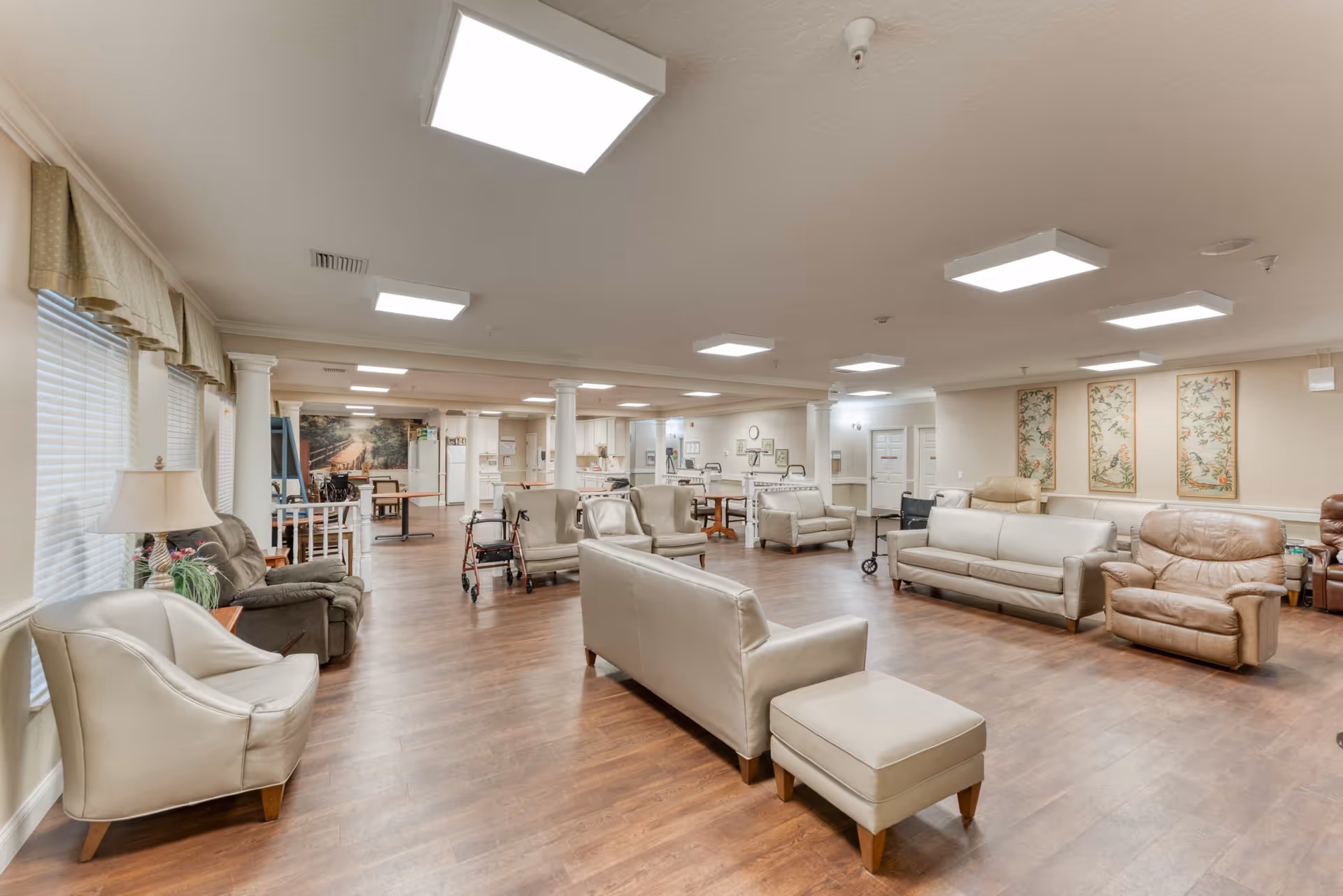 A spacious senior living facility common area with multiple beige and brown leather sofas and armchairs arranged on a wooden floor. The room is well-lit with ceiling lights and has large windows with blinds and valances on the left side. There are white columns, a few walkers, tables, and framed artwork on the walls. The background shows a kitchen area with white cabinets and appliances.