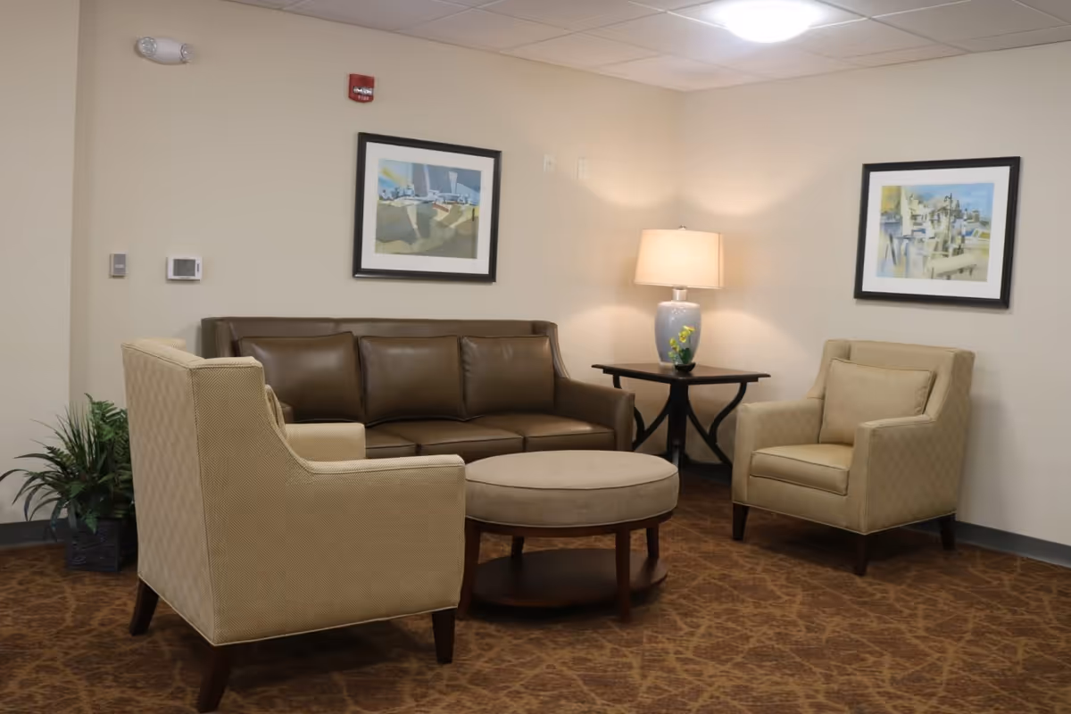 A cozy seating area in a senior living facility with a brown leather sofa, two beige upholstered armchairs, a round beige ottoman, a dark wooden side table with a lamp, two framed abstract paintings on the walls, and a potted plant in the corner.