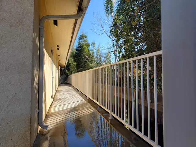 A narrow outdoor walkway alongside a building with a beige exterior wall on the left and a white metal railing on the right. The walkway is wet and reflects the railing and nearby trees. Trees and blue sky are visible beyond the railing.