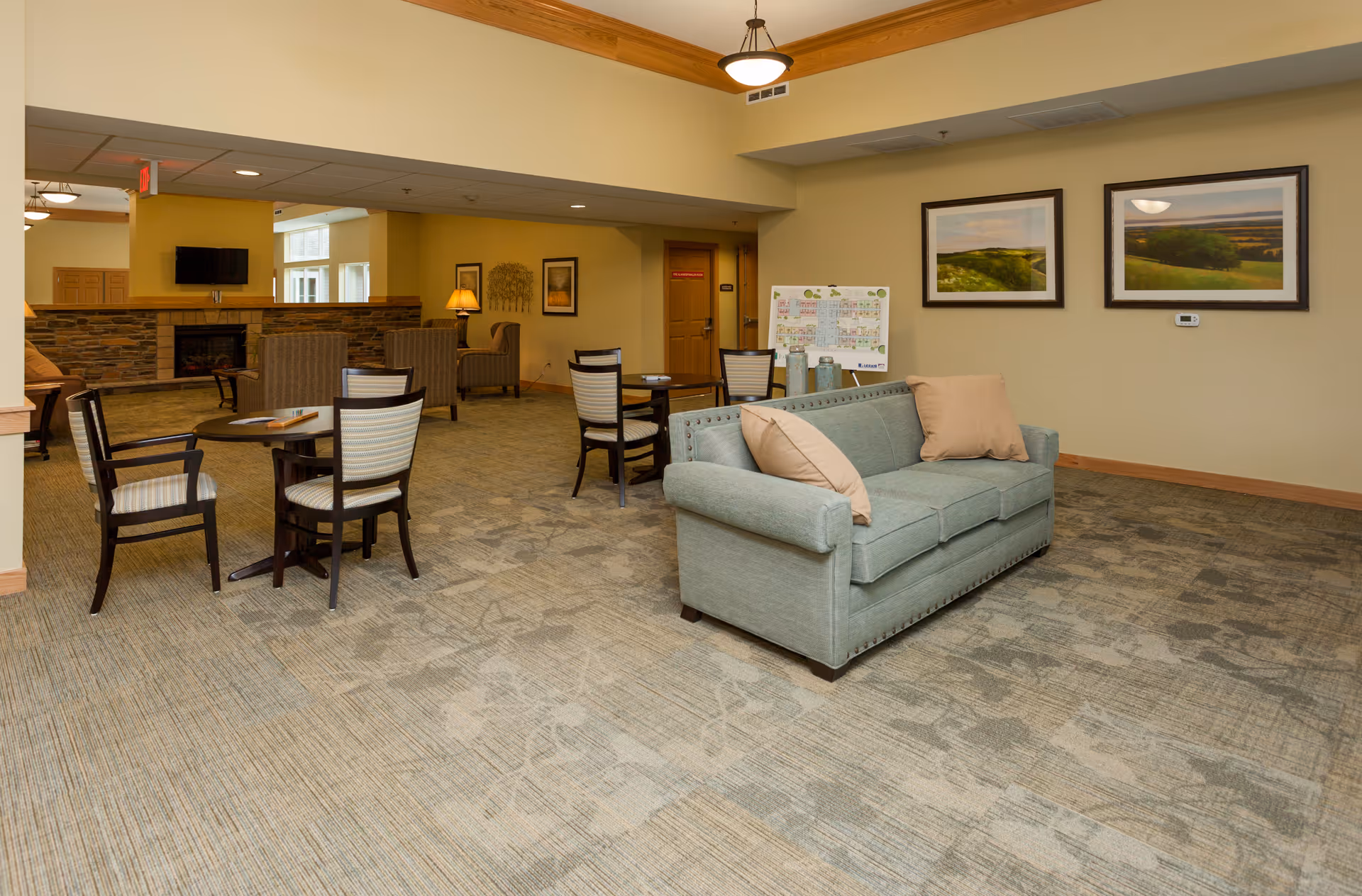 A spacious senior living common area with a light blue sofa with beige pillows, several wooden chairs with striped cushions around small round tables, a stone fireplace with a mounted TV above it, framed landscape paintings on the wall, and warm yellow lighting.