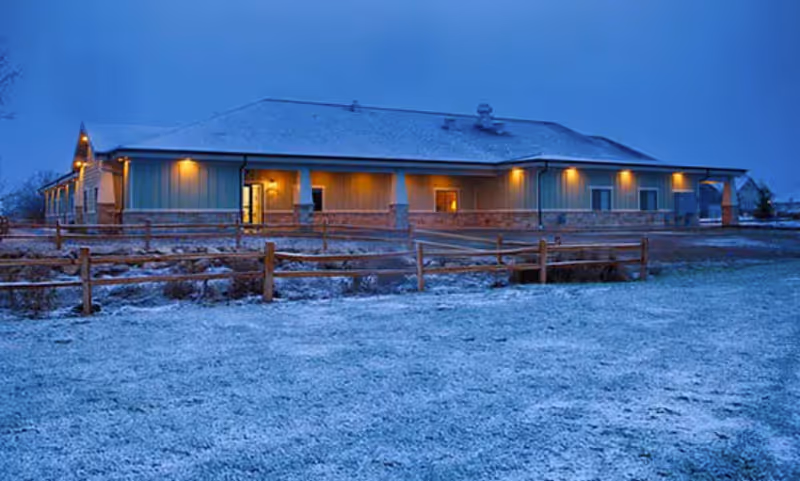 Exterior view of a single-story assisted living facility building at dusk with lights on and a light dusting of snow on the ground and roof. A wooden fence runs along the front of the property.