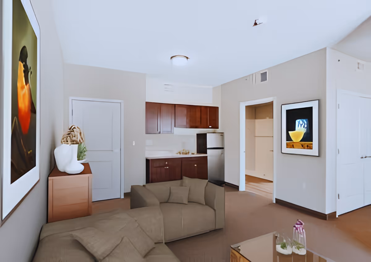 Interior view of a living area in an assisted living facility featuring a beige sectional sofa, a wooden cabinet with decorative items, a small kitchen with dark wooden cabinets and a stainless steel refrigerator, and two framed pictures on the walls. The room has beige walls and carpeted flooring.