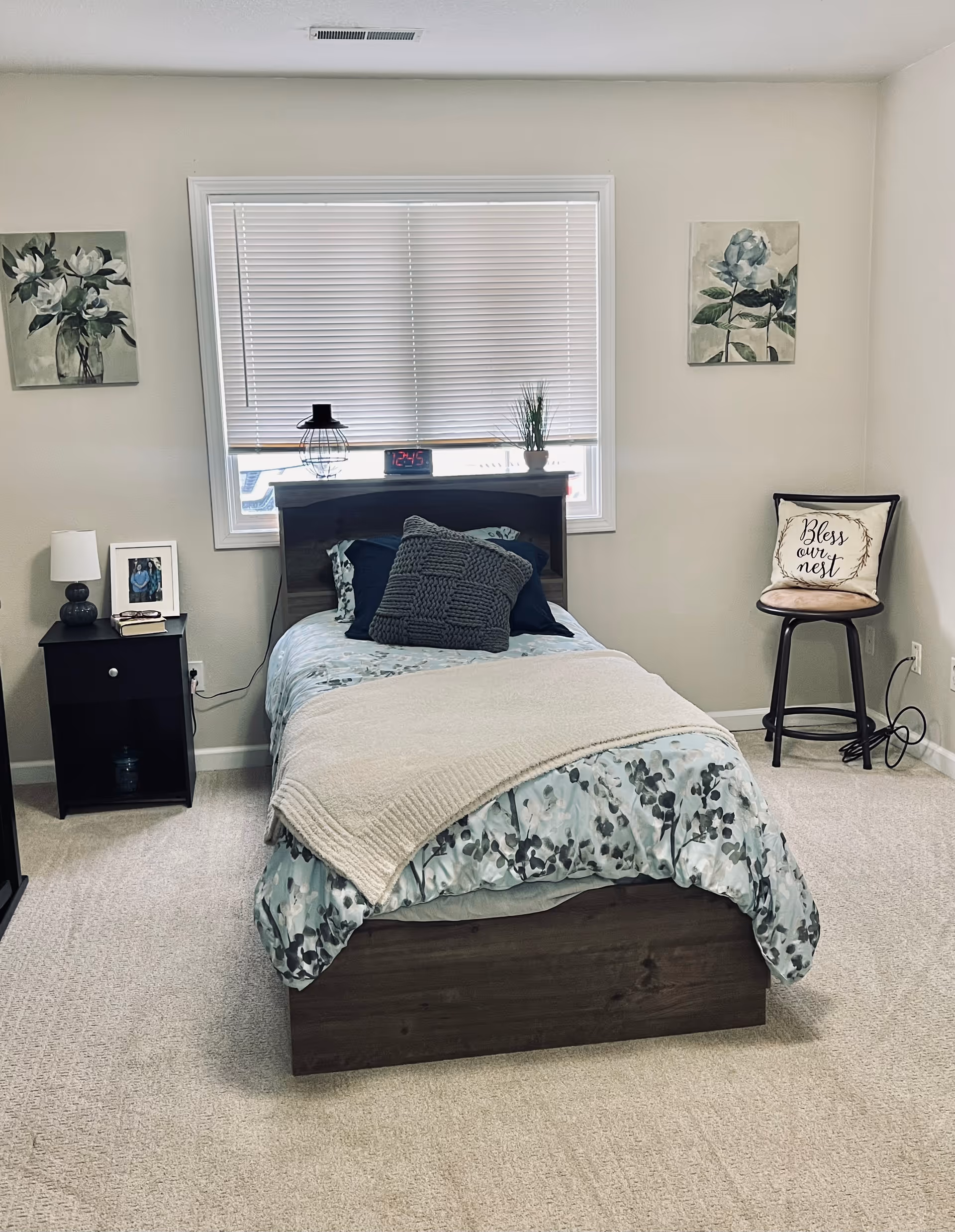A neatly made single bed with floral bedding and a beige throw blanket in a bedroom. The bed has a wooden headboard with a digital clock, a small lamp, and a potted plant on top. To the left of the bed is a black nightstand with a lamp, a framed photo, and a book. On the right side of the room, there is a tall stool with a decorative pillow that says 'Bless our nest'. The walls are light-colored with two floral paintings hanging on either side of the window behind the bed. The floor is carpeted in a light beige color.