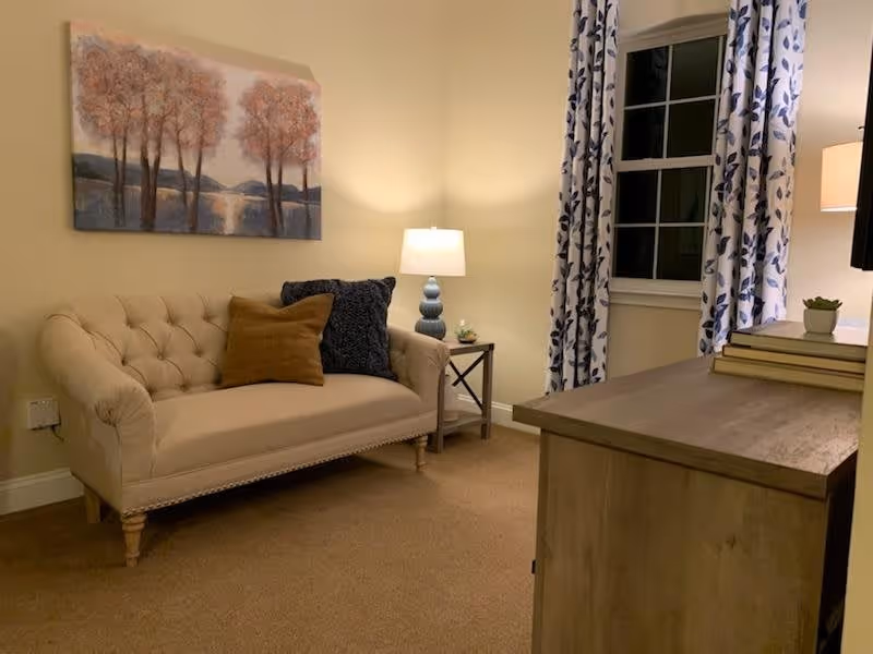 A cozy living room area with a beige tufted loveseat adorned with two pillows, one brown and one dark blue. A side table with a blue lamp and a small plant sits next to the loveseat. There is a window with white curtains featuring a blue leaf pattern. A wooden dresser with a stack of books and a small plant is visible on the right side. A painting of trees and a lake hangs on the wall above the loveseat.
