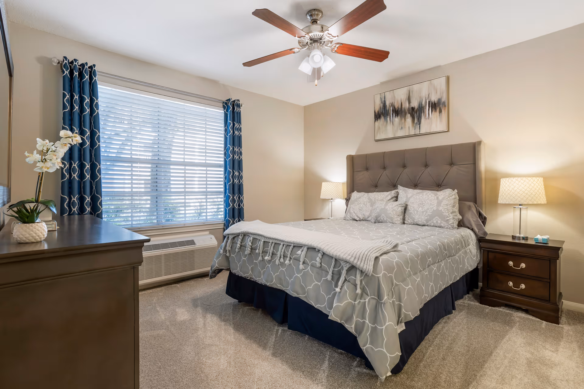 A neatly made bed with a tufted headboard in a bedroom. The bed has gray and white patterned bedding with pillows and a throw blanket. There are two wooden nightstands on either side of the bed, each with a lamp. A large window with blue patterned curtains and white blinds is on the left wall. A ceiling fan with wooden blades and lights is mounted on the ceiling. A dresser with a potted plant and decorative items is visible on the left side of the image.