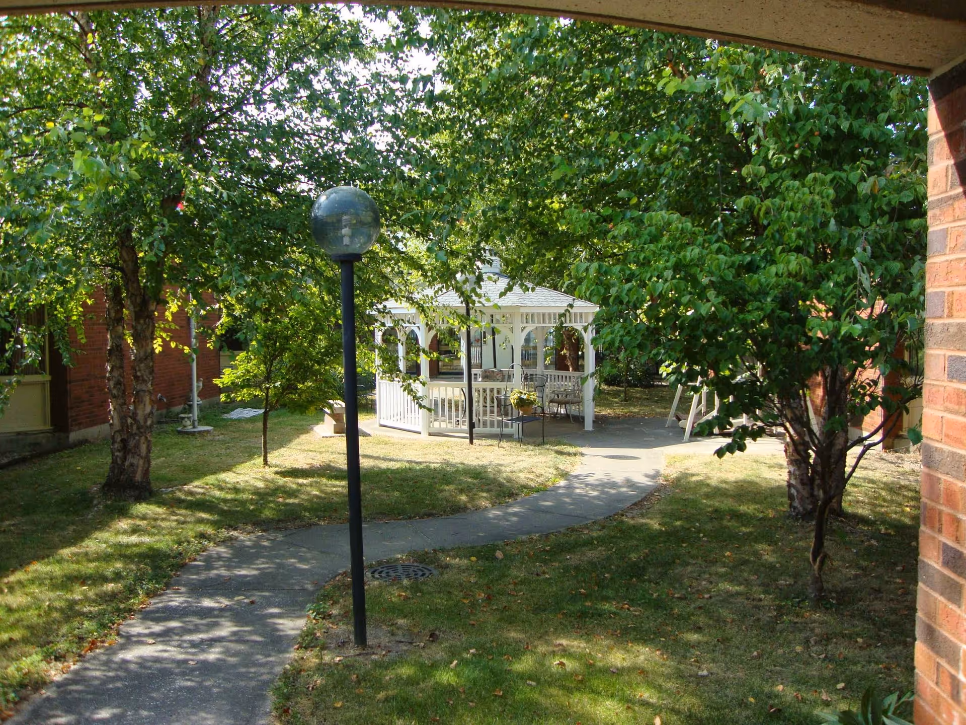 A garden area with a curved concrete pathway leading to a white gazebo surrounded by green trees and grass. There is a black lamp post along the path and brick buildings partially visible on the sides.