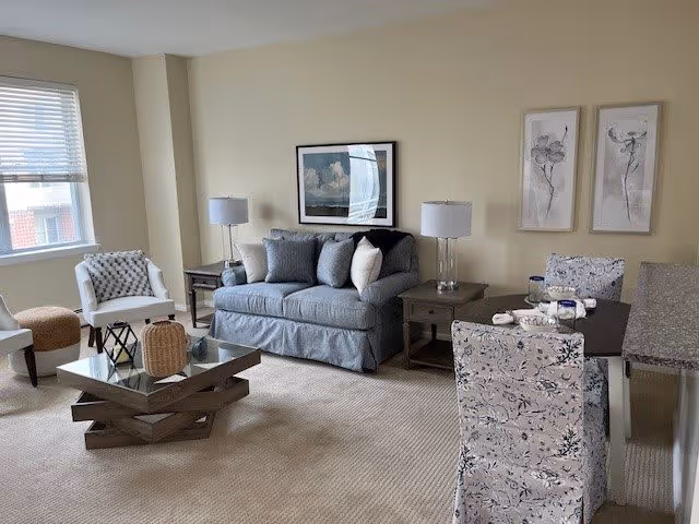 A cozy living room with a gray sofa adorned with multiple cushions, two matching side tables with lamps, a glass coffee table with decorative items, a white armchair near a window with blinds, and a dining area with floral-patterned chairs and a dark wooden table. The walls are decorated with framed artwork.