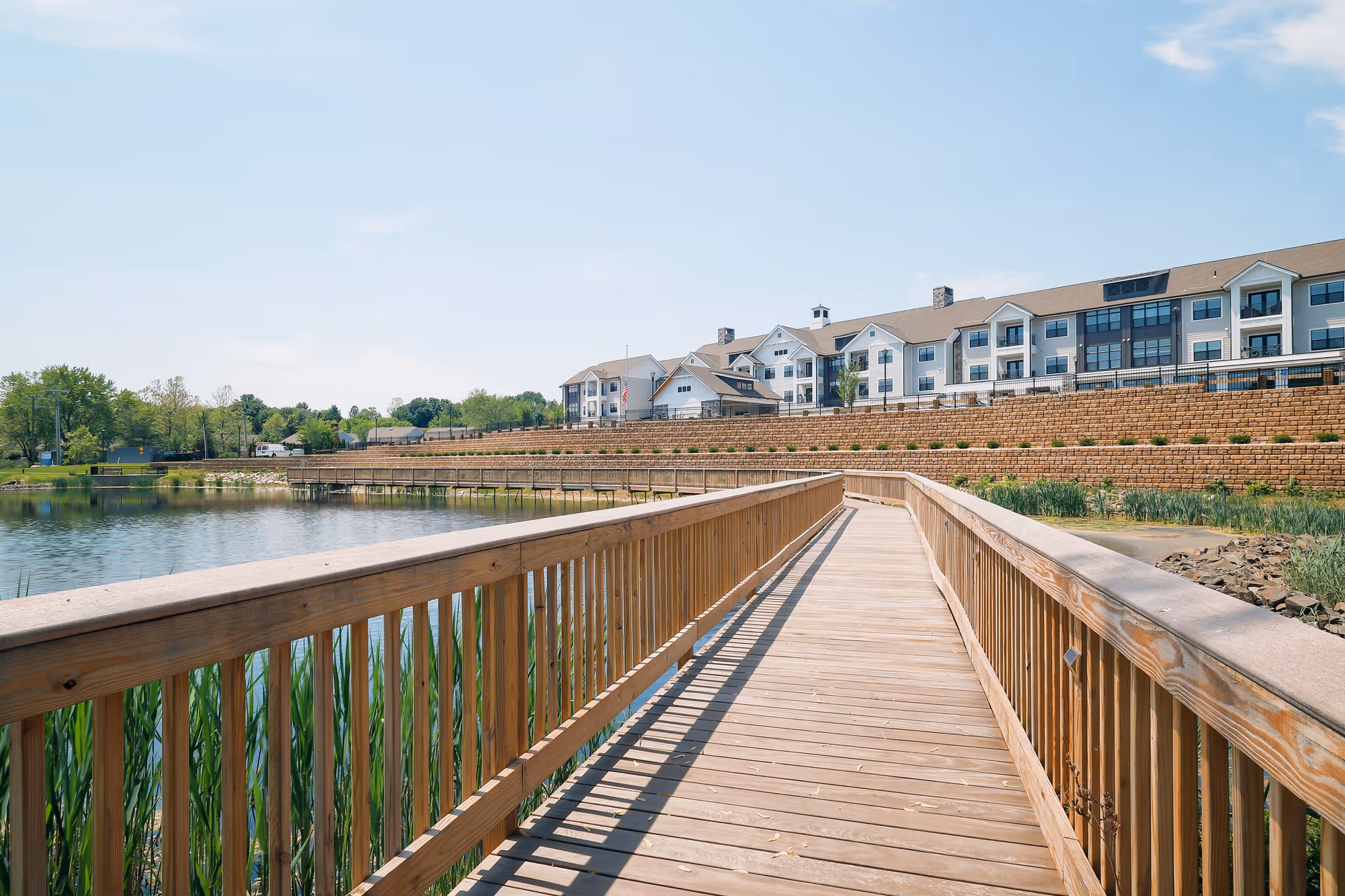 A wooden boardwalk extending over a pond with green reeds along the water's edge, leading towards a large multi-story residential building with white siding and multiple windows under a clear blue sky.