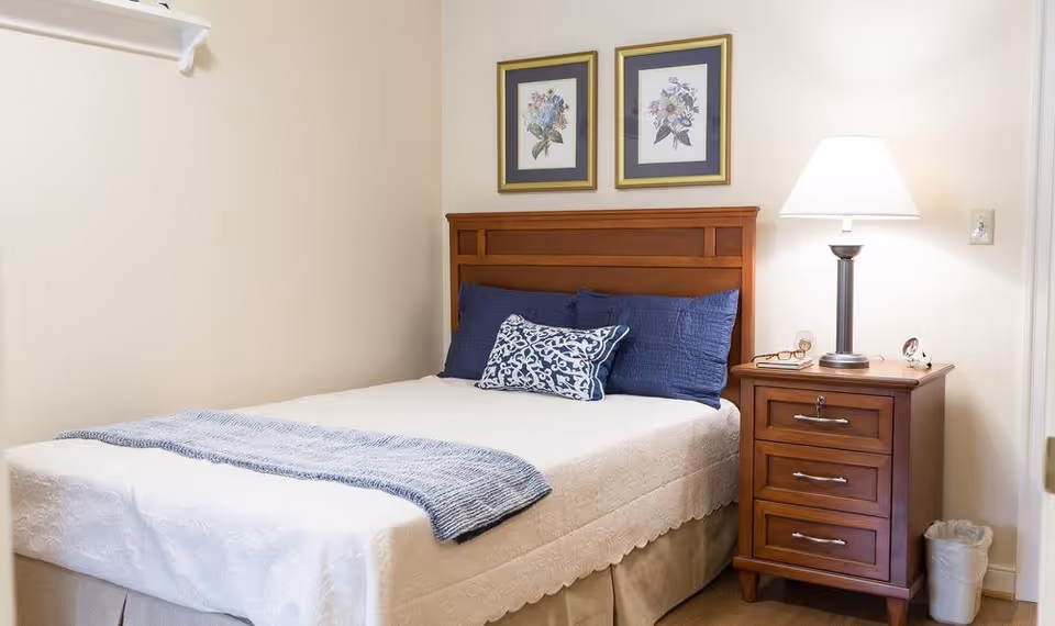 A neatly made bed with white bedding and blue pillows in a senior living bedroom. There is a wooden headboard and a matching wooden nightstand with a lamp, glasses, and a small clock. Two framed floral pictures hang on the wall above the bed.